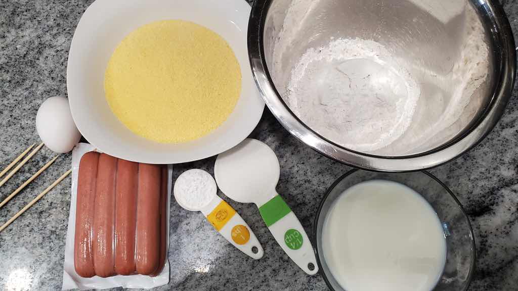 The ingredients to make air fryer corn dogs are corn meal, flour, egg, milk, hot dogs, baking powder, sugar and salt.