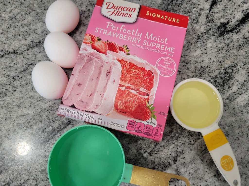 The ingredients needed are strawberry cake mix, eggs, oil, water, white candy melts, cream cheese, vanilla extract, powdered sugar, heavy cream and sprinkles.